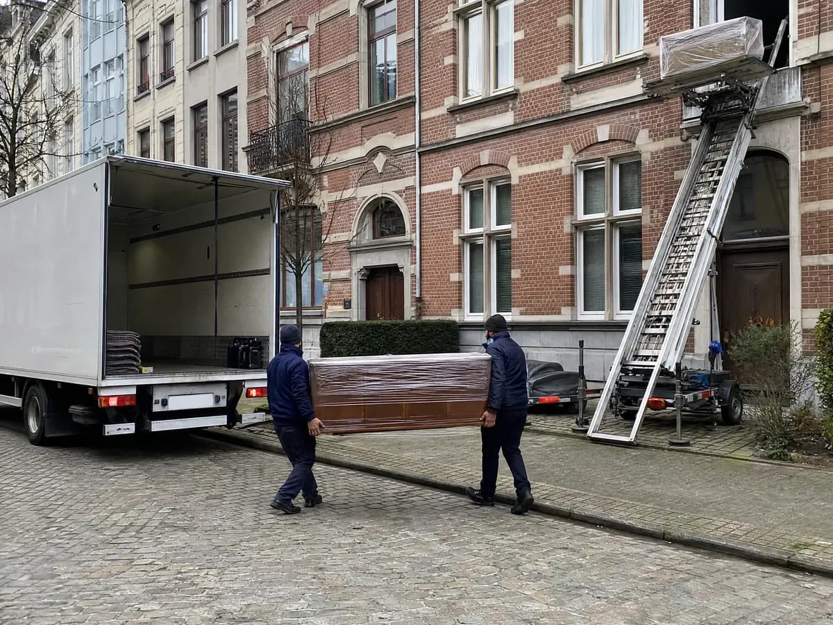 Antwerp movers carrying a wrapped dresser to the moving van, with the furniture lift against the facade
