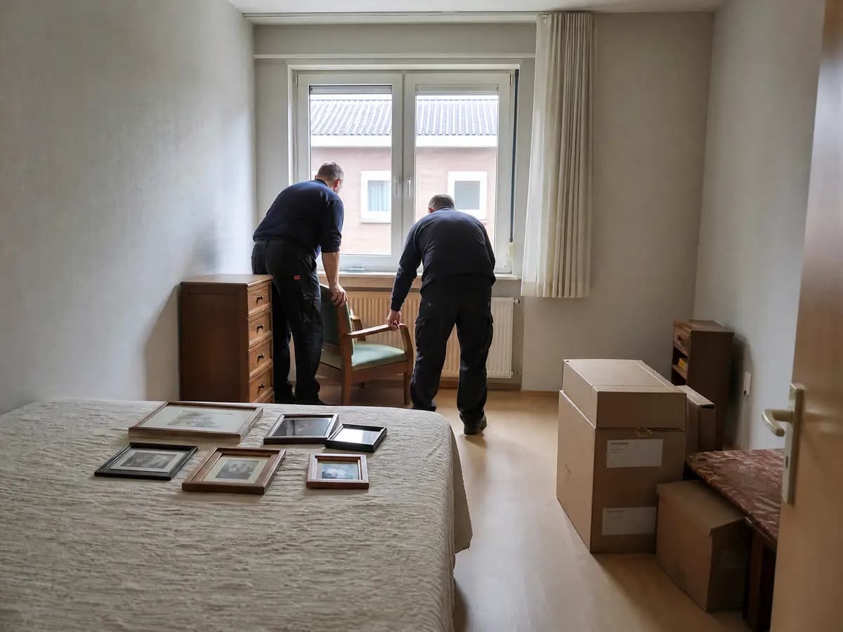Twee verhuizers zetten een groene fauteuil bij het raam van een slaapkamer, met ingelijste foto's op het bed