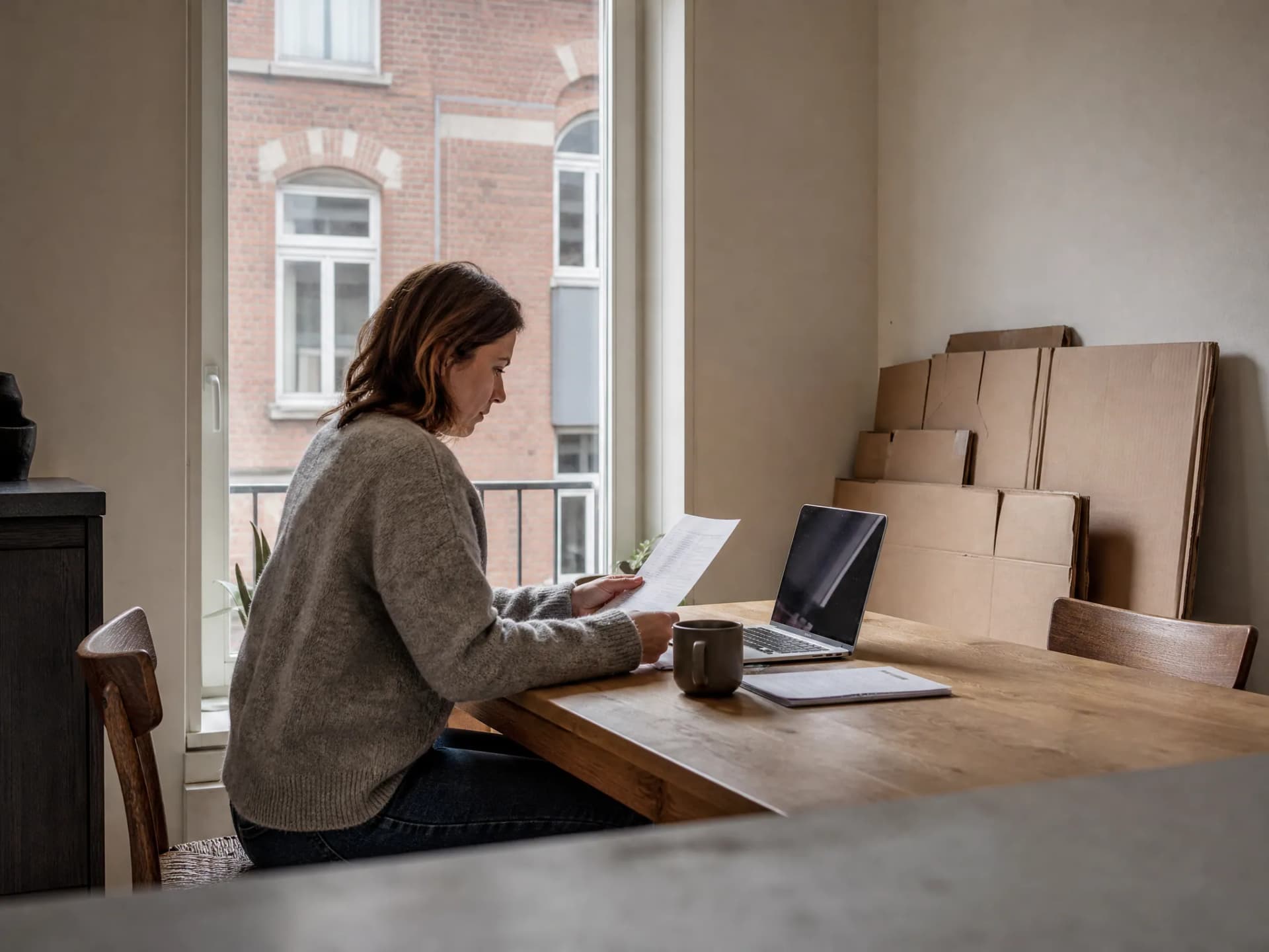 Een vrouw leest een verhuisofferte aan de keukentafel naast haar laptop, met platte verhuisdozen tegen de muur