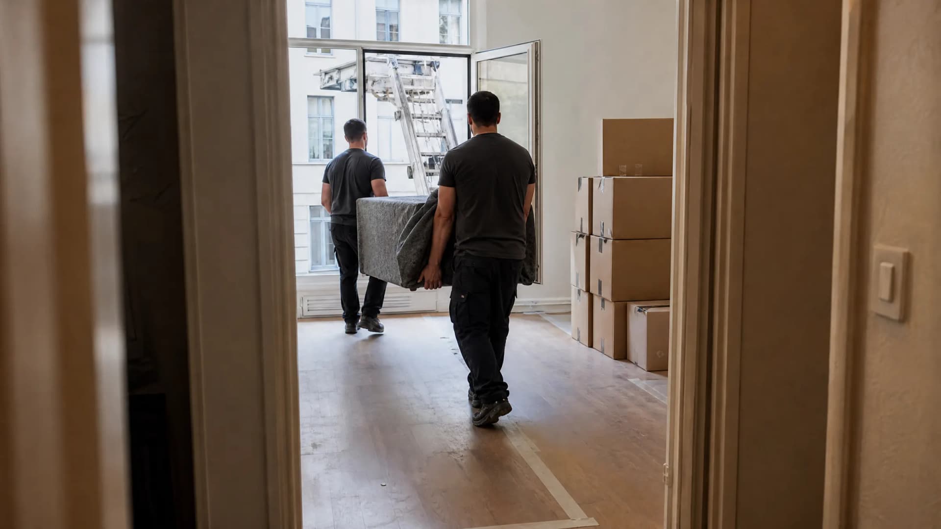 Twee verhuizers dragen een ingepakt meubel de lege woonkamer van een Antwerps appartement in; door het raam staat de ladderlift tegen de gevel