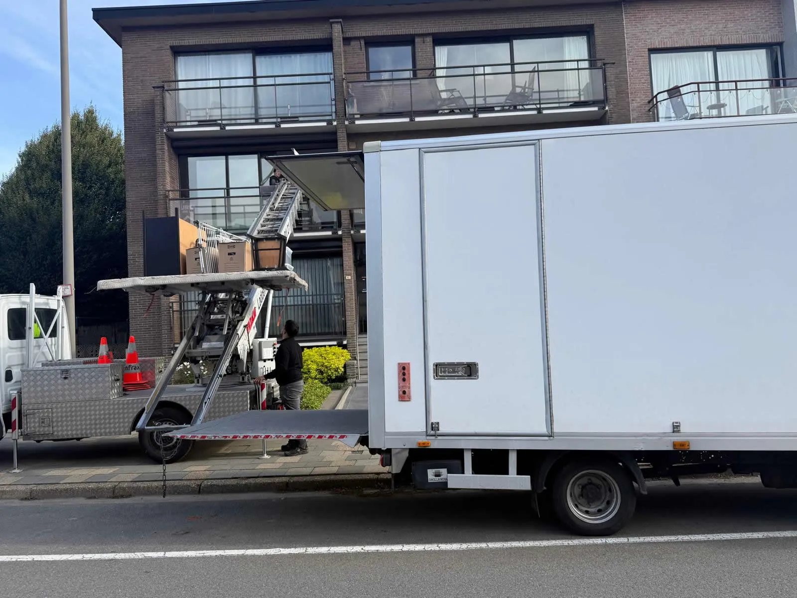 Verhuizen in Zwijndrecht: de ladderlift brengt een kast, dozen en een lamp naar het balkon van een appartementsgebouw, naast onze verhuiswagen met neergelaten laadklep; een verhuizer bedient de lift